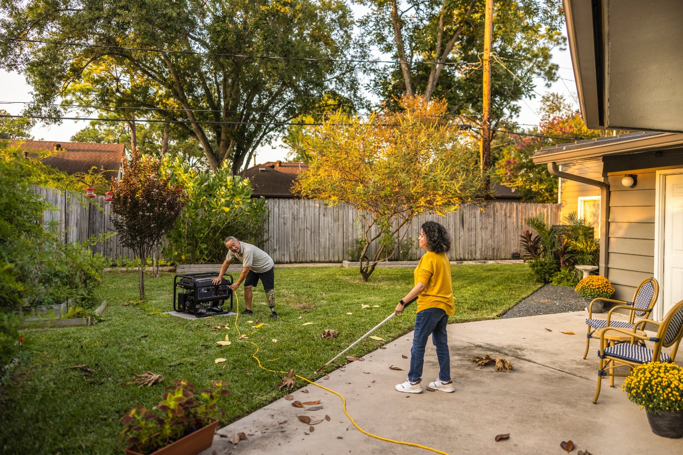 Portable generator running on a lawn away from a house while people manage an outdoor power connection.