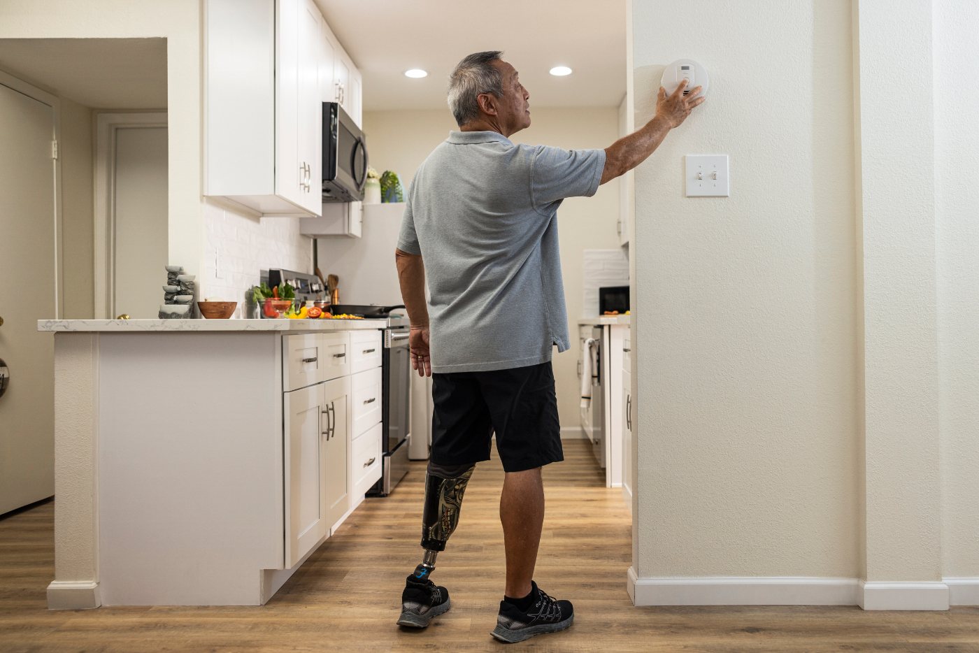 A man in a kitchen reaches up to test a smoke alarm mounted on an interior wall.