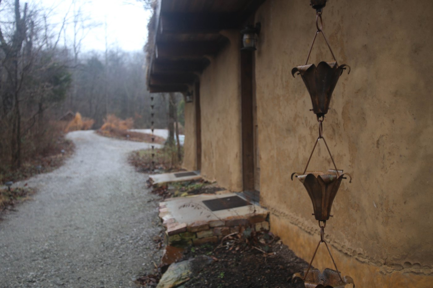 House-wall downspout beside a gravel path and rainwater collection containers in Makanda, Illinois.