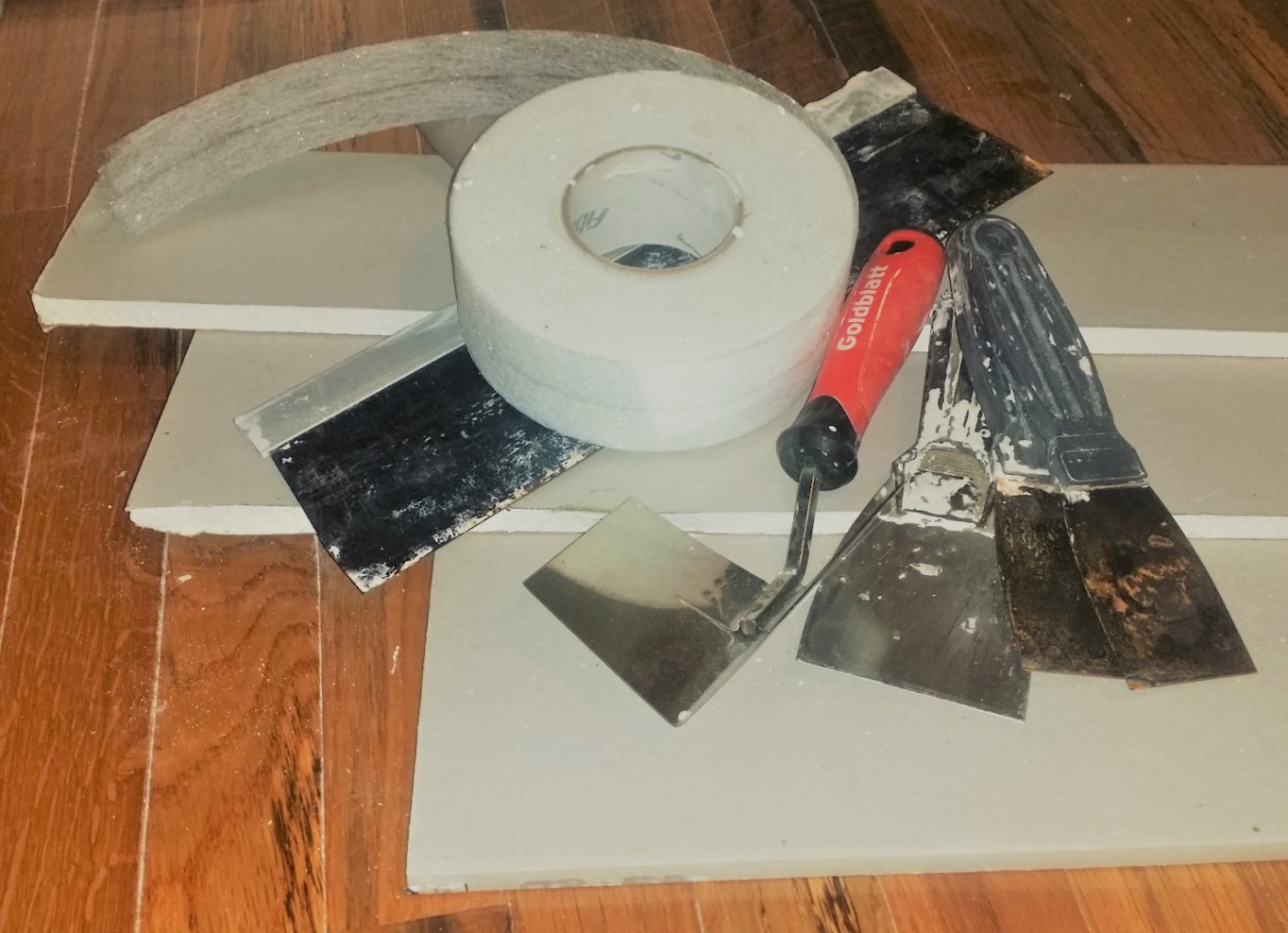 Cut drywall panels, joint tape, and drywall finishing tools arranged on a wood floor.